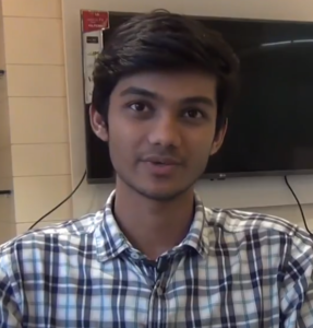 A young man with short dark hair, possibly an entrepreneur like Pranav Panpalia, is wearing a blue and white plaid shirt. He is sitting indoors in front of a television mounted on a wall. The background also includes a small section of tiled wall. He is looking slightly to his left.