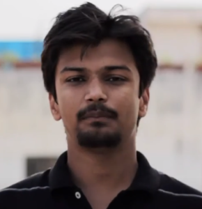 A man with dark hair and a beard is standing outdoors, looking directly at the camera. He is wearing a black collared shirt. The background is blurry and appears to be an urban setting, possibly hinting at the bustling environment where Ashish Sangai navigates his entrepreneurial journey.
