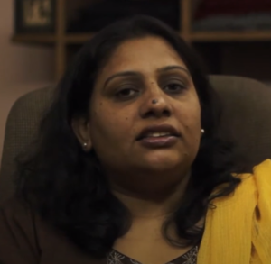 A woman with long dark hair is seated indoors. She is wearing a brown top with a yellow scarf draped over her left shoulder and small earrings. Shelves with folded clothes or books are visible in the background. She appears to be speaking or addressing the camera, possibly discussing Damyantii's market reach.