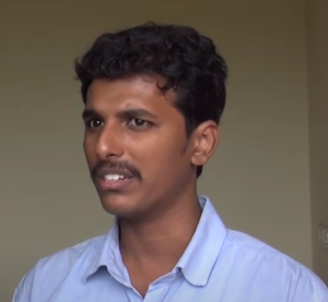 A young man with short, curly hair and a mustache is standing indoors. Wearing a light blue button-up shirt, he appears to be discussing business impact. He looks slightly to the side, with a plain, light-colored wall behind him, possibly during an NEN Mentoring session by the Wadhwani Foundation.