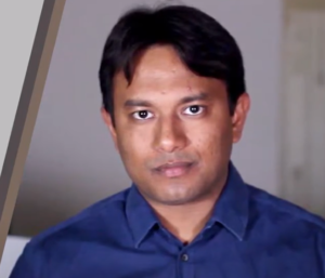 A man with short dark hair, wearing a blue button-up shirt, looks directly at the camera. The background is blurred, and there are diagonal gray and brown lines on the left side of the image. The photograph exudes a professional aura that suggests he might be discussing entrepreneurial resources from the Wadhwani Foundation.