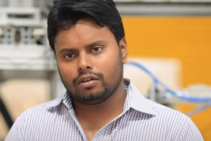 A man with short dark hair and a beard, wearing a striped collared shirt, looks past the camera. The background is blurred, with a glimpse of machinery from Nikit Engineers and a yellow wall.