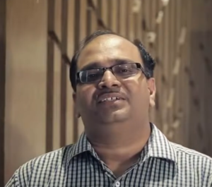 A man wearing glasses and a checkered shirt smiles at the camera, standing in front of a background featuring vertical wooden panels. He appears confident and ready with tools for growth, benefiting from initiatives like those by the Wadhwani Foundation.