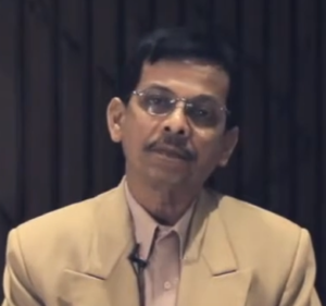 A man with short dark hair and glasses, wearing a beige suit jacket over a light pink shirt, is looking directly at the camera. He appears to be in a formal setting with a dark striped background. Representing the Wadhwani Foundation, a microphone can be seen clipped to his jacket.