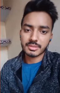 A young man with dark hair and a beard is looking at the camera. He is wearing a blue shirt and a black jacket, with earphones in his ears. He appears to be indoors, with shelves and some items visible in the background, embodying the spirit of NEN NextGen entrepreneurship in a bustling Digital City.