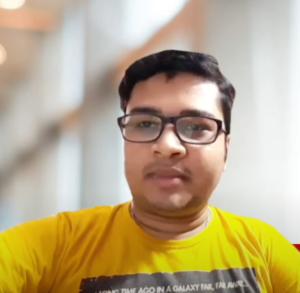 A man with short dark hair and glasses is wearing a yellow shirt with text on it. He is indoors, and the background is blurred, showing structures that might be part of a building or room. He appears to be looking slightly off-camera, perhaps contemplating his next big idea for a student venture.