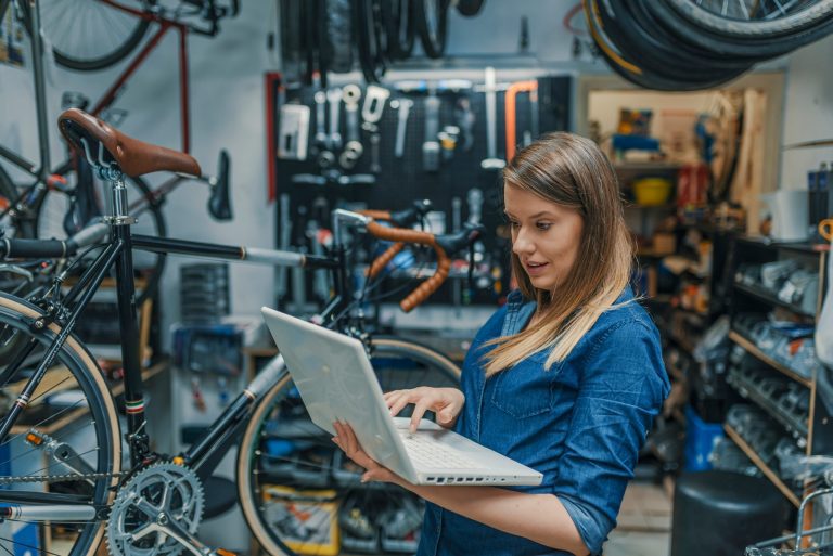 A focused woman in a denim shirt using a laptop in a well-organized bicycle repair shop, with various tools and bike parts in the background, enhances her employability skills.