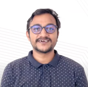 A person with short dark hair, glasses, and a beard smiles at the camera. They are wearing a dark polo shirt with a small, light-colored pattern. The background is white with subtle diagonal lines, embodying the spirit of the NEN IGNITE Program known for enhancing technical knowledge.