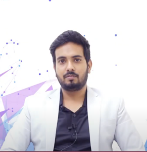 A man with a beard and mustache wearing a white blazer over a black shirt is looking directly at the camera. He has short dark hair and a neutral expression. The background features abstract purple and blue geometric shapes and dots, hinting at an NEN IGNITE event, with Palash Dhawale's perfect pitch evident as he has a microphone clipped to his shirt.