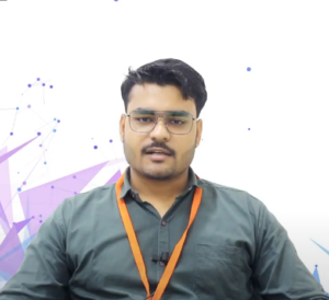 A man with short black hair, wearing glasses and dressed in a dark green shirt, sits against a white background with purple geometric and blue dot designs. He has an orange lanyard around his neck and a small microphone clipped to his shirt, preparing for his pitch before investors at the NEN IGNITE Program.