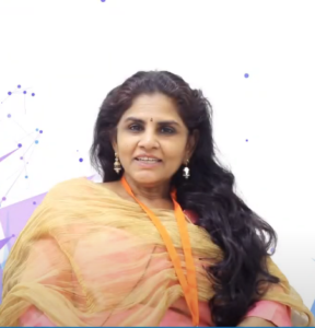 A woman with long, dark hair is smiling at the camera. She is wearing a light-colored shawl and an orange lanyard with a white background, which has light blue and purple abstract graphics. The lanyard signifies her participation in the NEN IGNITE program, identifying her as entrepreneur Pratibha Iyer.