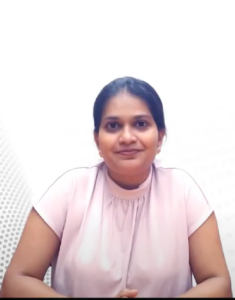 A woman with a calm expression sits against a light, dotted background. She has dark hair pulled back and wears a light pink short-sleeved blouse. Slightly leaning forward with her hands clasped, she seems deep in thought, perhaps reflecting on her experience in the InFED-NEN IGNITE Program.
