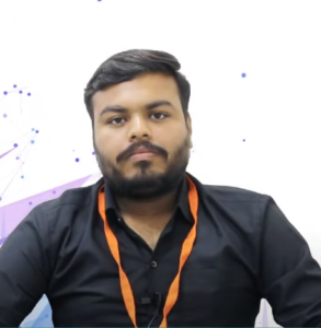 A man with short, dark hair and a beard is wearing a black shirt and an orange lanyard that reads "NEN IGNITE." He is sitting against a white background adorned with abstract blue and purple geometric shapes and dotted lines, embodying the innovative spirit of a startup. His expression is neutral as he looks directly forward.