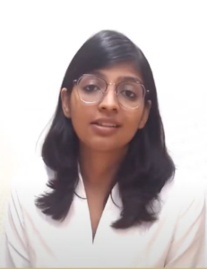 A person with shoulder-length black hair and glasses, wearing a white shirt, is looking towards the camera and speaking about the InFED-NEN IGNITE Program. The background is plain white.