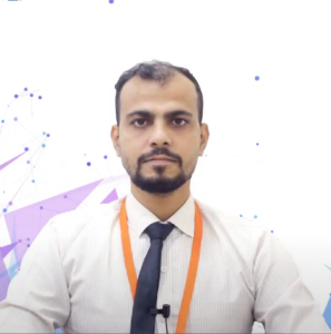 Shahbaz Ahmad Siddiqui, a man with a beard wearing a light-colored shirt, dark tie, and an orange lanyard, sits against a white background with abstract blue and purple geometric designs. A small microphone is clipped to his shirt as he looks directly at the camera, ready to discuss the NEN IGNITE Program.