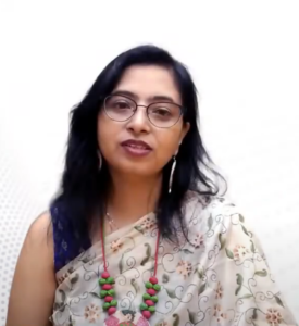 A woman with long black hair and glasses is wearing a floral saree and a beaded necklace. She is looking directly at the camera with a neutral, yet approachable expression. The plain, light-colored background highlights her participation in the InFED-NEN IGNITE Program.