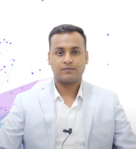 A person wearing a light gray blazer and white shirt, sitting against a white background with blue and purple abstract designs. They have short dark hair, are looking at the camera, and a small microphone is clipped to the shirt, giving the structured program an air of professionalism.