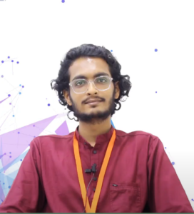 A person with curly hair, glasses, and a beard wearing a maroon shirt with an orange lanyard sits against a background of geometric and abstract blue and purple shapes. A microphone is clipped to their shirt. Siddharth Gupta addresses budding entrepreneurs at the NEN IGNITE Program.
