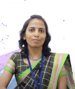A woman with shoulder-length dark hair is wearing a green and black sari with a traditional pattern. She is also wearing a blue lanyard, indicating her involvement in the NEN IGNITE Program, and has a subtle smile. The background is white with abstract dotted and linear designs.