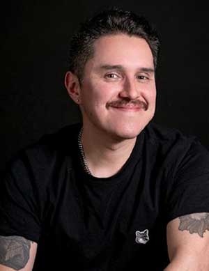 A smiling person with a mustache, wearing a black t-shirt with the EmpowerED México logo, with visible tattoos on both arms, against a dark background.