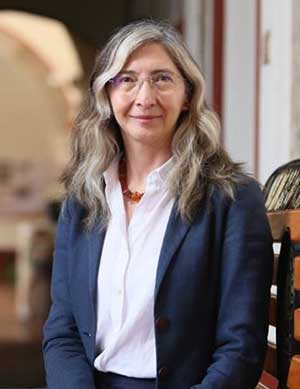 A professional woman with long, gray hair wearing glasses, a white blouse, and a dark blazer stands confidently indoors, with a partial view of an elegant wooden railing on the side and an archway