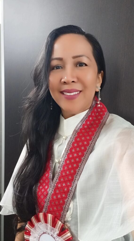 Uma mulher em trajes tradicionais com um xale vermelho e dourado sorri gentilmente em frente a uma porta de madeira, adornada com longos cabelos escuros e brincos de prata.