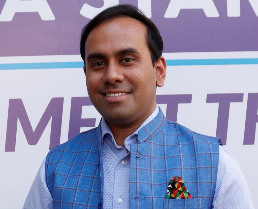 A smiling man wearing a blue checked waistcoat adorned with a colorful pocket square stands before a sign with the text "eastar meet the".