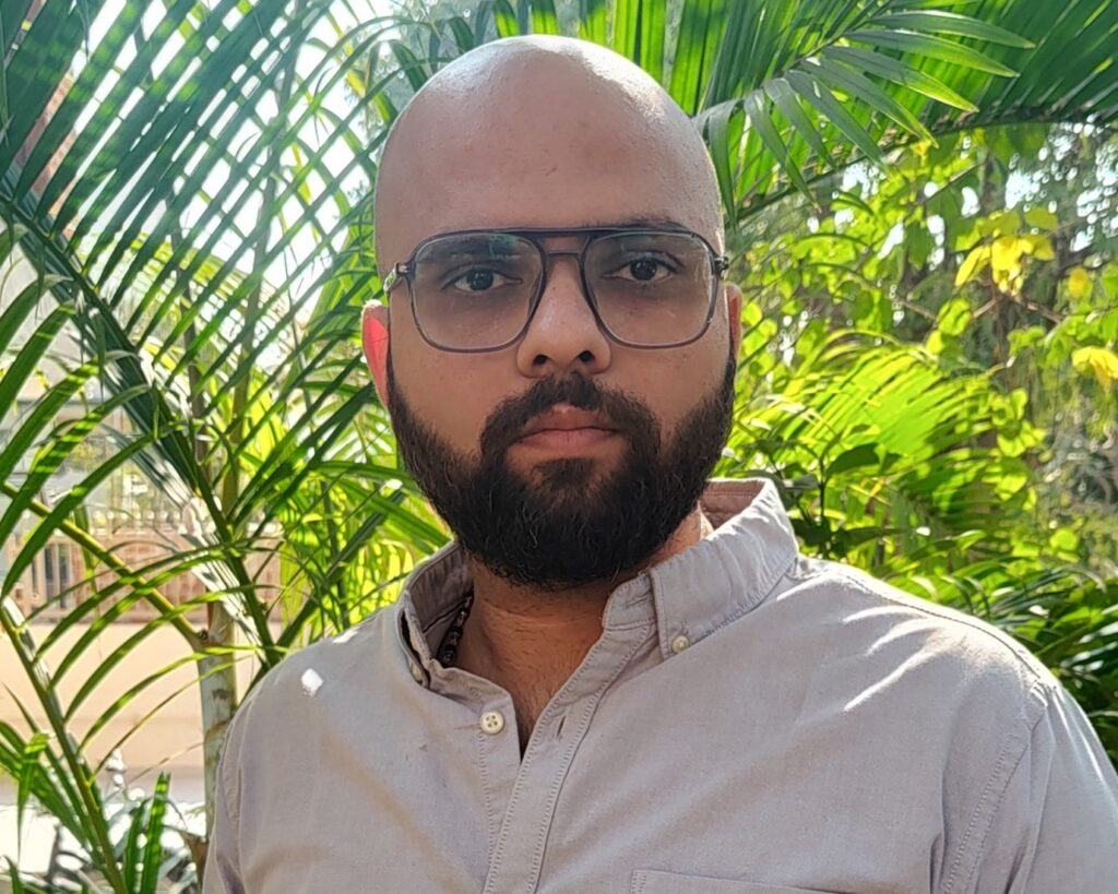 Um homem com barba e óculos em pé em frente a folhas verdes de palmeira, vestindo uma camisa cinza clara, olhando diretamente para a câmera.