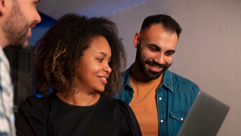 Dos colegas, una mujer y un hombre, sonríen mientras exploran juntos nuevas tecnologías en un ordenador portátil. La mujer tiene el pelo rizado y lleva un top negro, mientras que el hombre tiene barba y lleva un