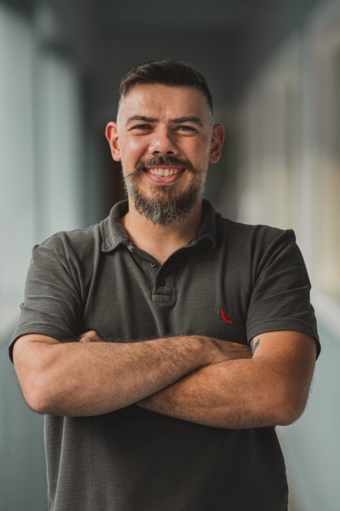 Um homem sorrindo para a câmera, de pé, com os braços cruzados. Ele tem barba e um corte de cabelo curto, vestindo uma camisa polo escura. o fundo está suavemente desfocado.
