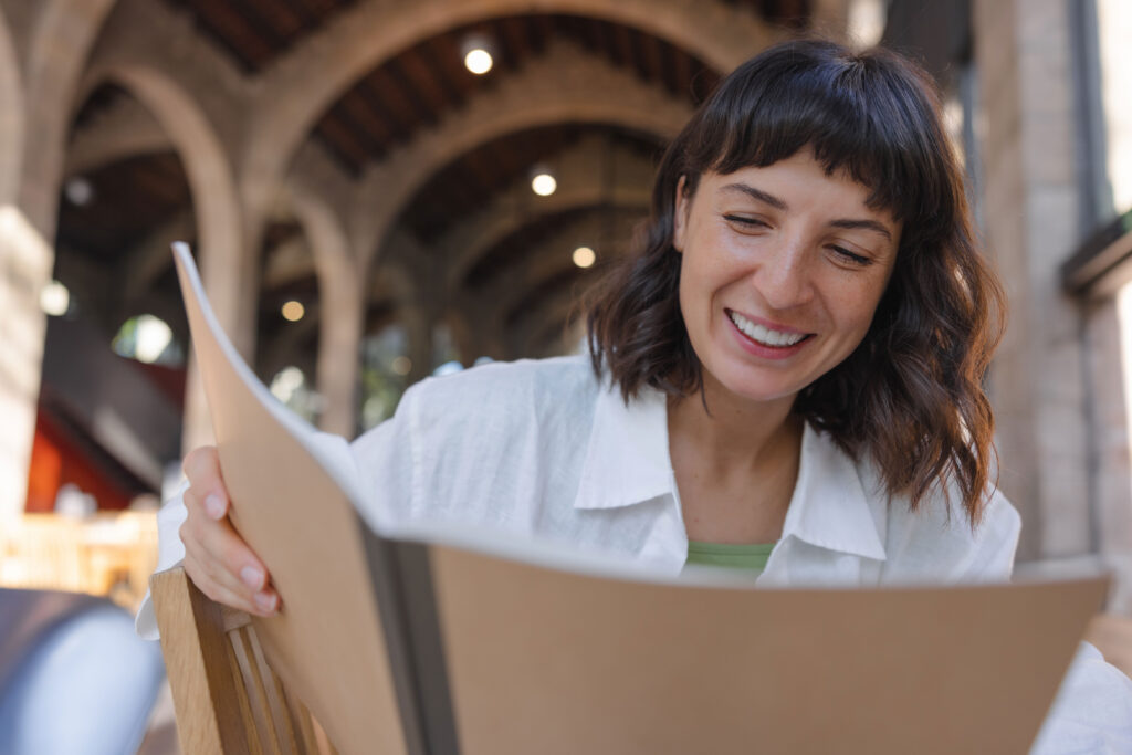 Una mujer sonriente con el pelo corto y castaño lee un libro sentada a una mesa de madera en una estructura arqueada con columnas, lo que sugiere un compromiso en un lugar público o al aire libre.