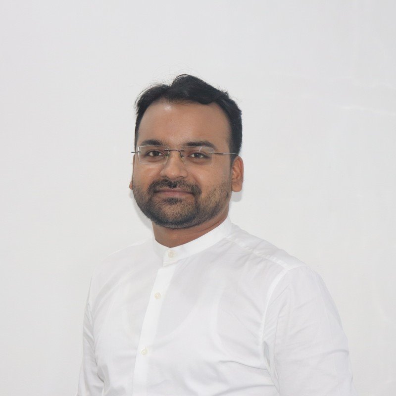 A man with short, dark hair, a beard, and glasses is smiling at the camera. He is wearing a white buttoned-up shirt against a plain white background.