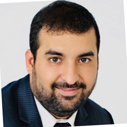 A close-up portrait of Rajnish Kapur, a man with a beard and short hair, wearing a dark suit and a white shirt with a dark tie. He has a slight smile and is looking directly at the camera. The background is plain and light-colored.