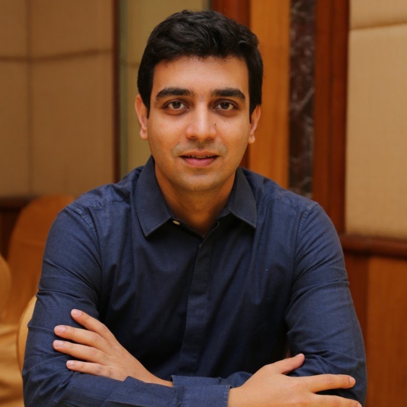 A person with short dark hair, wearing a dark blue long-sleeved shirt, sits with their arms crossed on a wooden table. They are in an indoor setting with wood-paneled and cream-colored walls in the background.