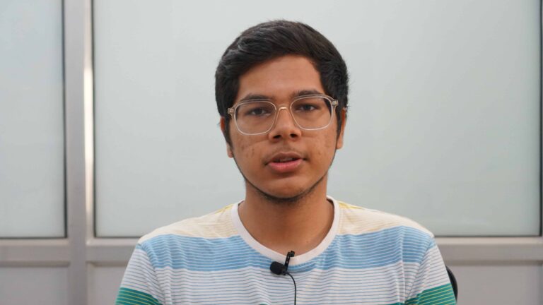 Adityaya Dhande, a person with short dark hair, glasses, and a trimmed beard, is wearing a striped shirt while sitting in front of a light-colored background. They have a serious expression and are wearing a microphone clipped to their shirt.