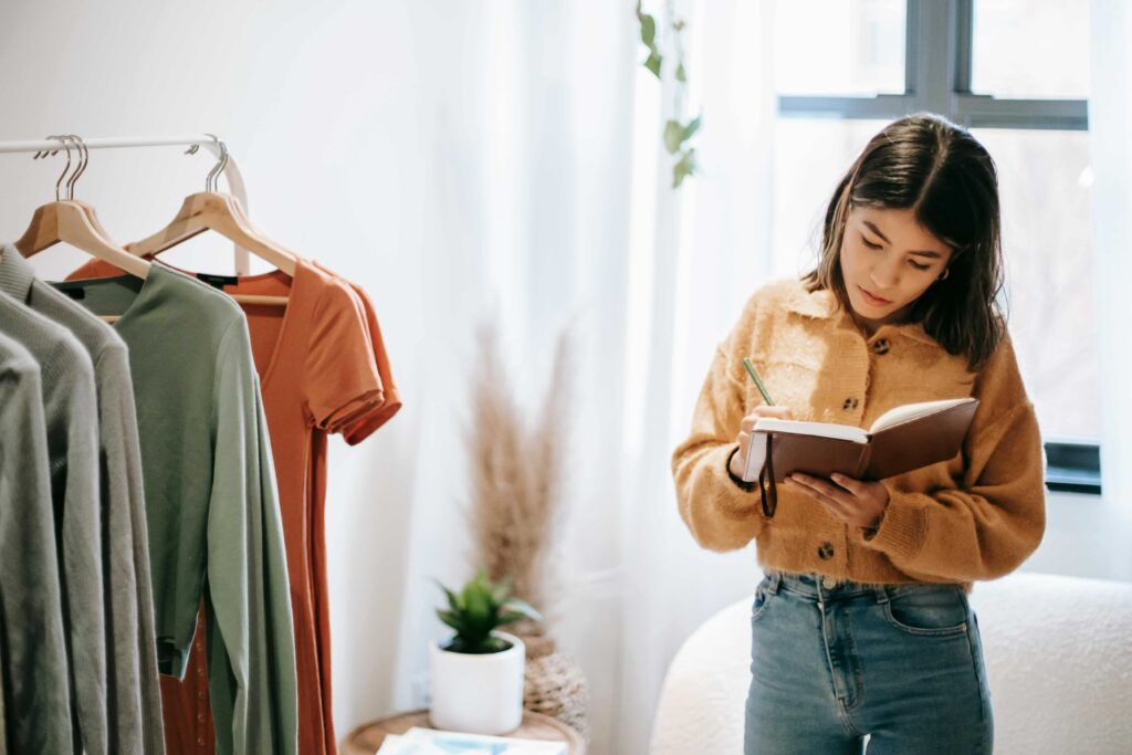 Seorang wanita dengan sweater coklat muda dan celana jins menulis di buku catatan sambil berdiri di samping rak berisi pakaian yang digantung. Ruangan yang dipenuhi cahaya alami dari jendela, tirai putih, dan pot tanaman di atas meja di dekatnya, mewujudkan suasana tenang yang ideal untuk merencanakan kesuksesan karier.