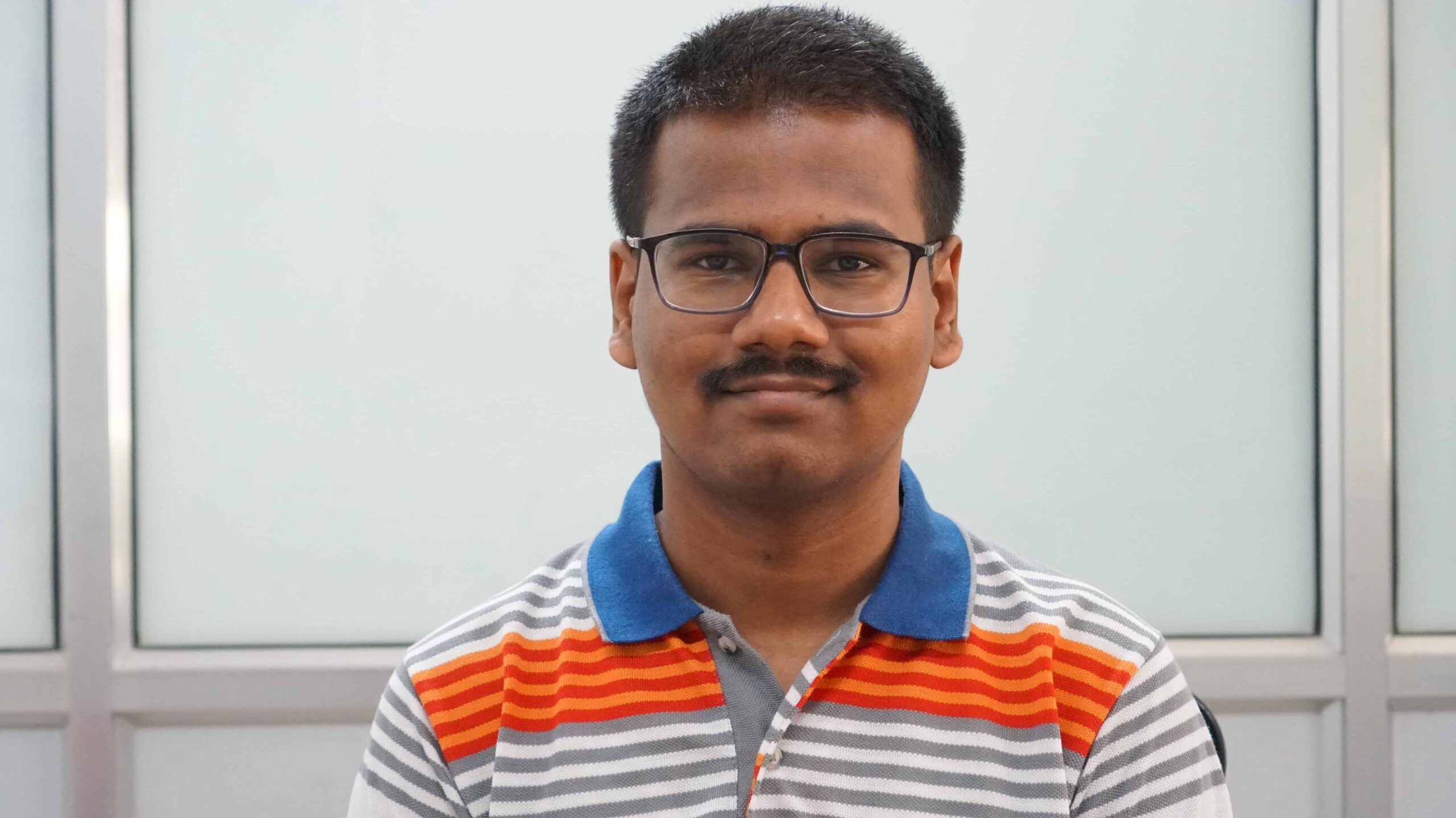 En la imagen aparece una persona con el pelo corto y negro, que lleva gafas y bigote. Harish Mokashi lleva una camisa de cuello con rayas horizontales de color rojo, naranja, gris y blanco. Detrás de él se ve un telón de fondo blanco y gris.
