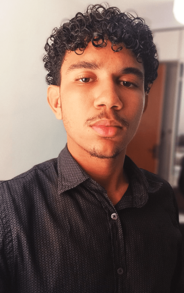 A young man with short, curly hair and a light mustache, named João Victor Alves, is looking at the camera with a neutral expression. He is wearing a dark, buttoned shirt and standing indoors with a door slightly ajar in the background.