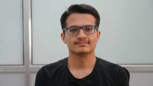 Sahil Wani, seorang pemuda berambut hitam pendek dan berkacamata berbingkai biru, menghadap ke arah kamera dan sedikit tersenyum. Dia mengenakan kaus hitam. Latar belakangnya menampilkan panel kaca buram dan bingkai metalik.