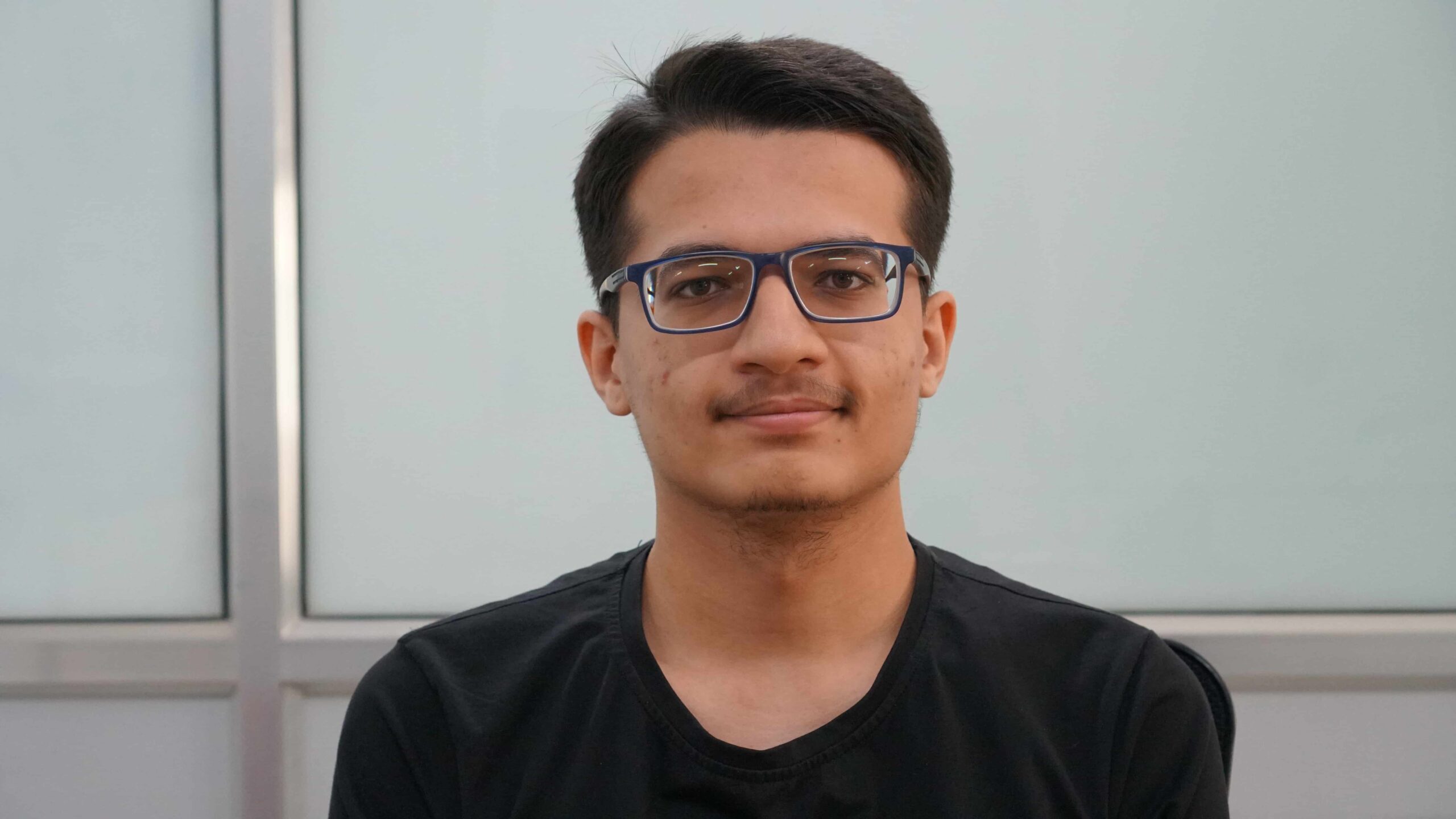 Sahil Wani, un joven con el pelo corto y oscuro y gafas de montura azul, mira a la cámara y sonríe ligeramente. Lleva una camiseta negra. El fondo presenta paneles de vidrio esmerilado y un marco metálico.