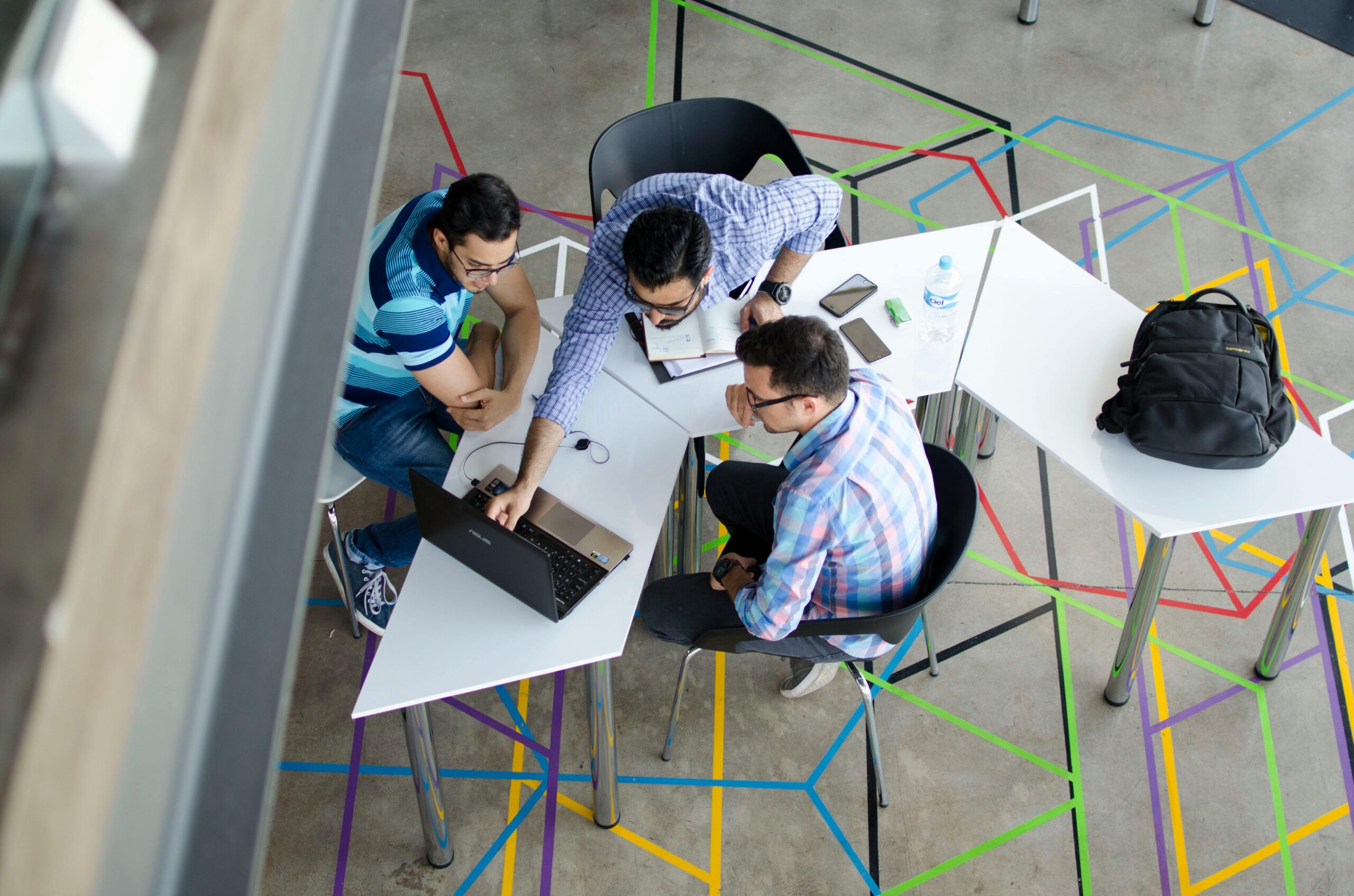 Three people are seated around a white table, collaborating on an innovative project. One person is pointing at a laptop screen, another is writing in a notebook, and a third is listening intently. The floor boasts colorful geometric designs, and there are additional tables and chairs nearby to foster networking.